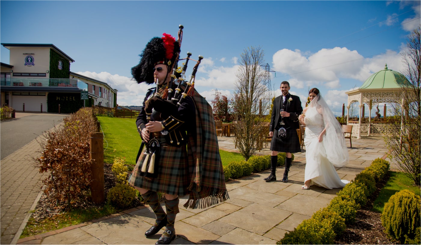 Medium wide shot of piper, Philip Barlow, leading an outdoor wedding processional